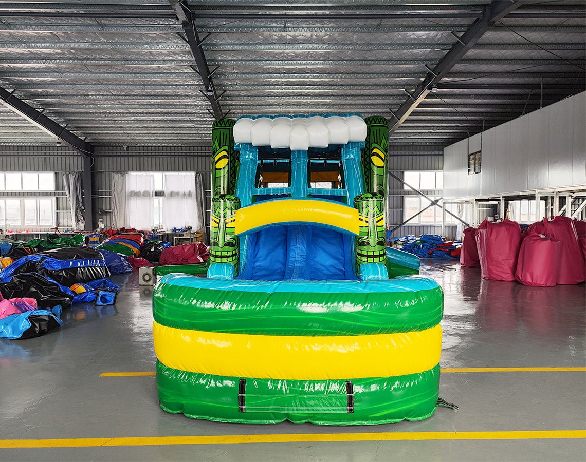 Front view of Tiki Wave Dual Lane Commercial Inflatable Combo set up in a large indoor warehouse. The inflatable features green tiki-style pillars, dual slides in blue and yellow, and a blue wave-like top with white inflatable foam. A green and yellow striped splash pool is visible at the base. Various other inflatable products and equipment are visible in the background.