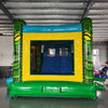 Rear view of Tiki Wave Dual Lane Commercial Inflatable Combo showing the entrance area. Features green tiki-style pillars with face designs, a yellow bounce house frame with mesh windows, and a blue top. The inflatable is set up on a green base. Various other inflatable products and equipment are visible in the warehouse background.