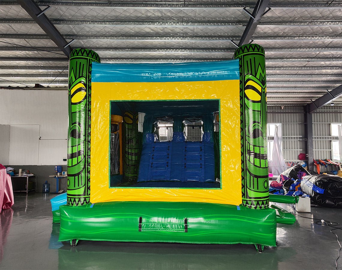 Rear view of Tiki Wave Dual Lane Commercial Inflatable Combo showing the entrance area. Features green tiki-style pillars with face designs, a yellow bounce house frame with mesh windows, and a blue top. The inflatable is set up on a green base. Various other inflatable products and equipment are visible in the warehouse background.