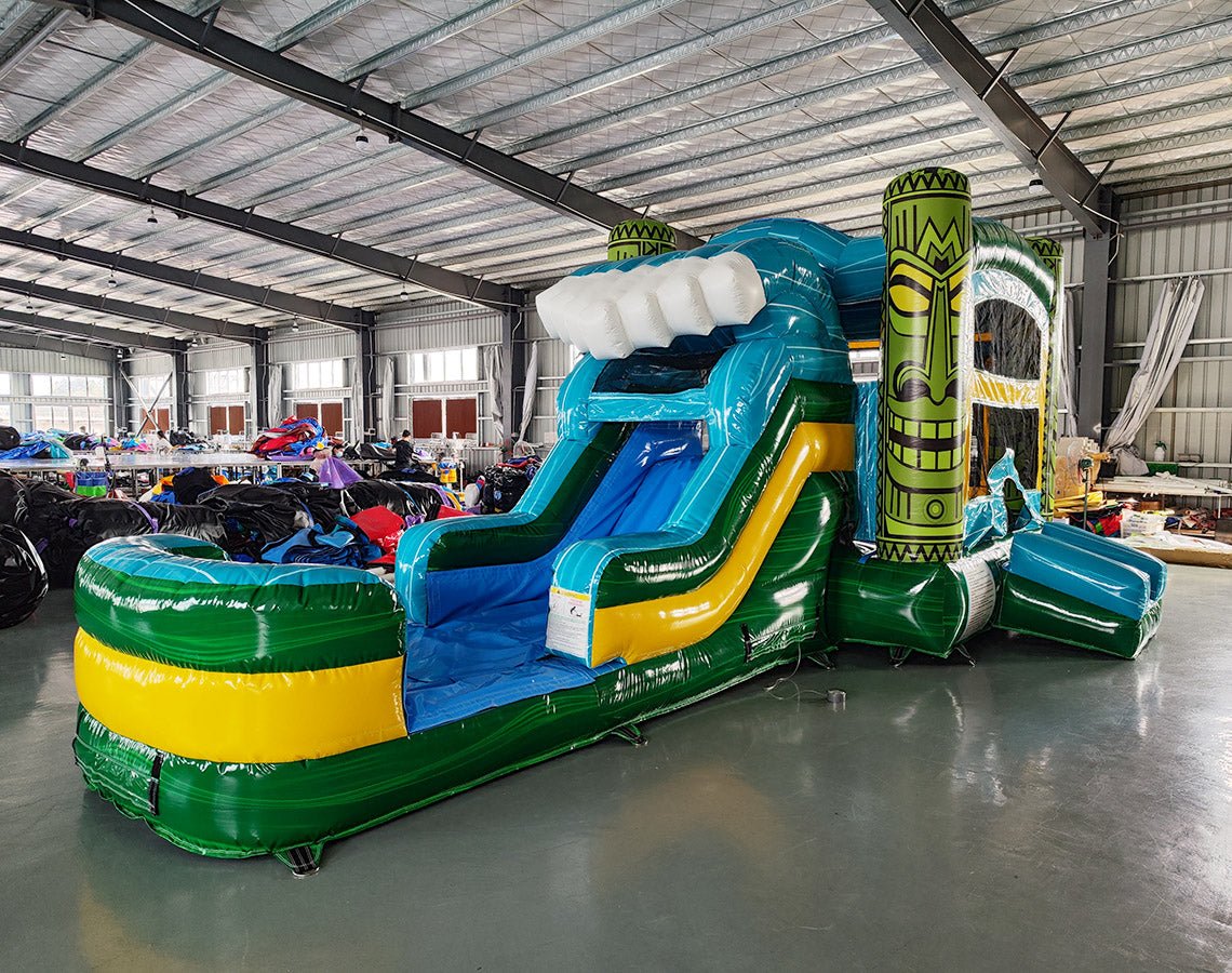 Side view of Tiki Wave Commercial Econo Inflatable Combo (Wet/Dry) showcasing the large blue curved slide with white inflatable wave crest, yellow bounce house structure, and green tiki face pillars. The combo includes a splash pool area and is set against a white background, highlighting its tropical theme and wet/dry functionality.