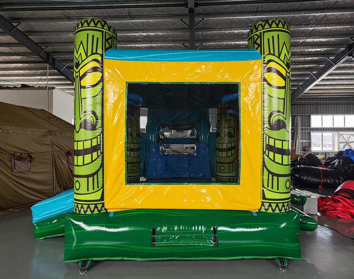 Rear view of Tiki Wave Commercial Econo Inflatable Combo (Wet/Dry) featuring a vibrant yellow bounce house with green tiki face pillars, blue top, and mesh entrance. Includes a curved blue slide, inflatable steps, and a green base. Set up in an indoor warehouse with other inflatables visible in the background.