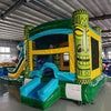 Wide-angle view of Tiki Wave Commercial Econo Inflatable Combo (Wet/Dry) in an indoor warehouse setting. The inflatable features a yellow bounce house with green tiki face pillars, blue curved slide with white wave crest, and multiple play areas. Safety instructions visible on the front. Other inflatable products and warehouse equipment visible in the background.