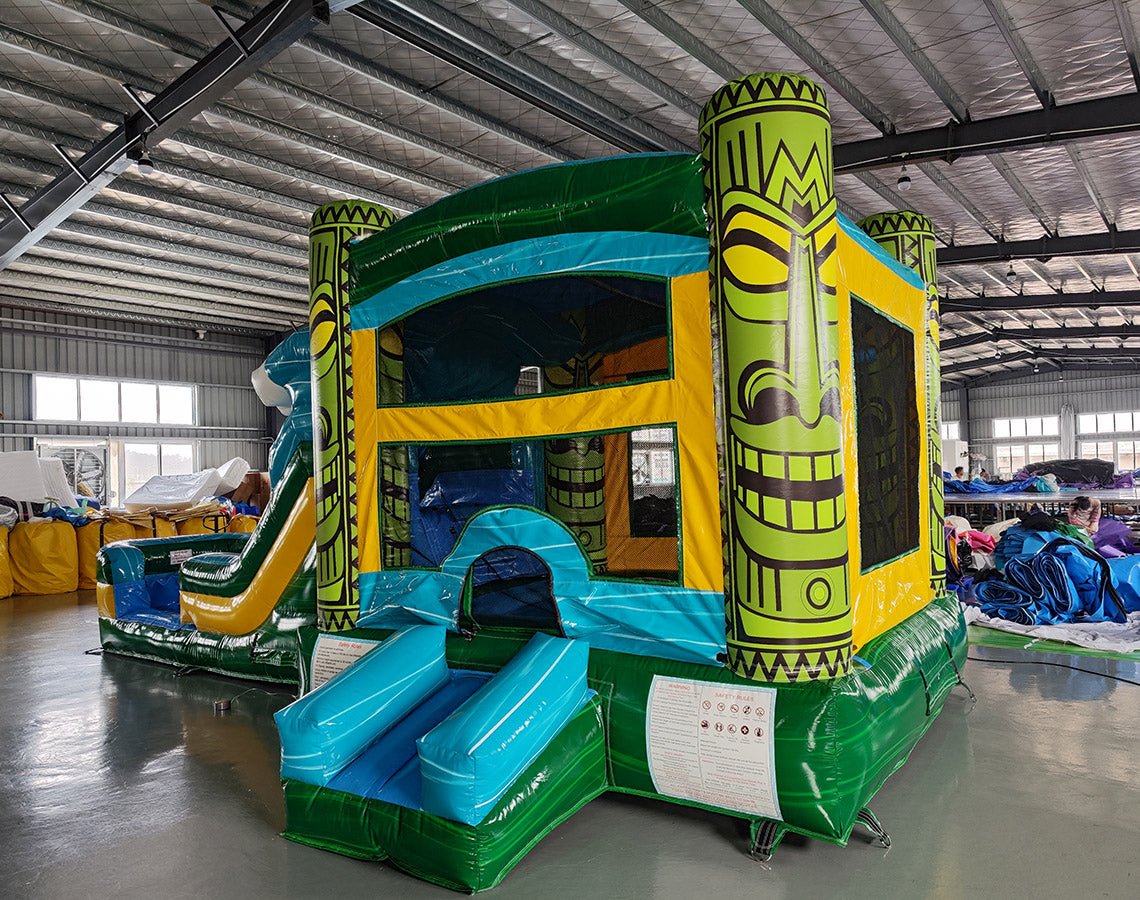 Wide-angle view of Tiki Wave Commercial Econo Inflatable Combo (Wet/Dry) in an indoor warehouse setting. The inflatable features a yellow bounce house with green tiki face pillars, blue curved slide with white wave crest, and multiple play areas. Safety instructions visible on the front. Other inflatable products and warehouse equipment visible in the background.