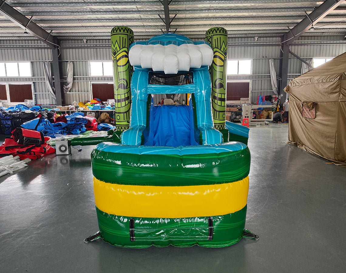 Front view of Tiki Wave Commercial Econo Inflatable Combo (Wet/Dry) featuring a vibrant tropical-themed design. The inflatable structure includes a large blue curved slide with white foam crest, yellow bounce house with green tiki face pillars, and a green base with yellow stripe. An inflatable cloud and palm tree top the structure. The combo is set up in an indoor warehouse, with other inflatables and equipment visible in the background, showcasing its commercial use and wet/dry functionality.