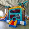 Side view of Tropic Shock Dual Lane Commercial Inflatable Combo (Wet/Dry) showcasing its full length. The inflatable features brown palm tree pillars with turquoise leaves, dual red slides, a turquoise frame, and a colorful base with blue, yellow, and red sections. A large circular splash pool is visible at one end, while the other end shows a yellow bounce house entrance. Set up in a spacious warehouse with metal roofing and various other inflatable products in the background.