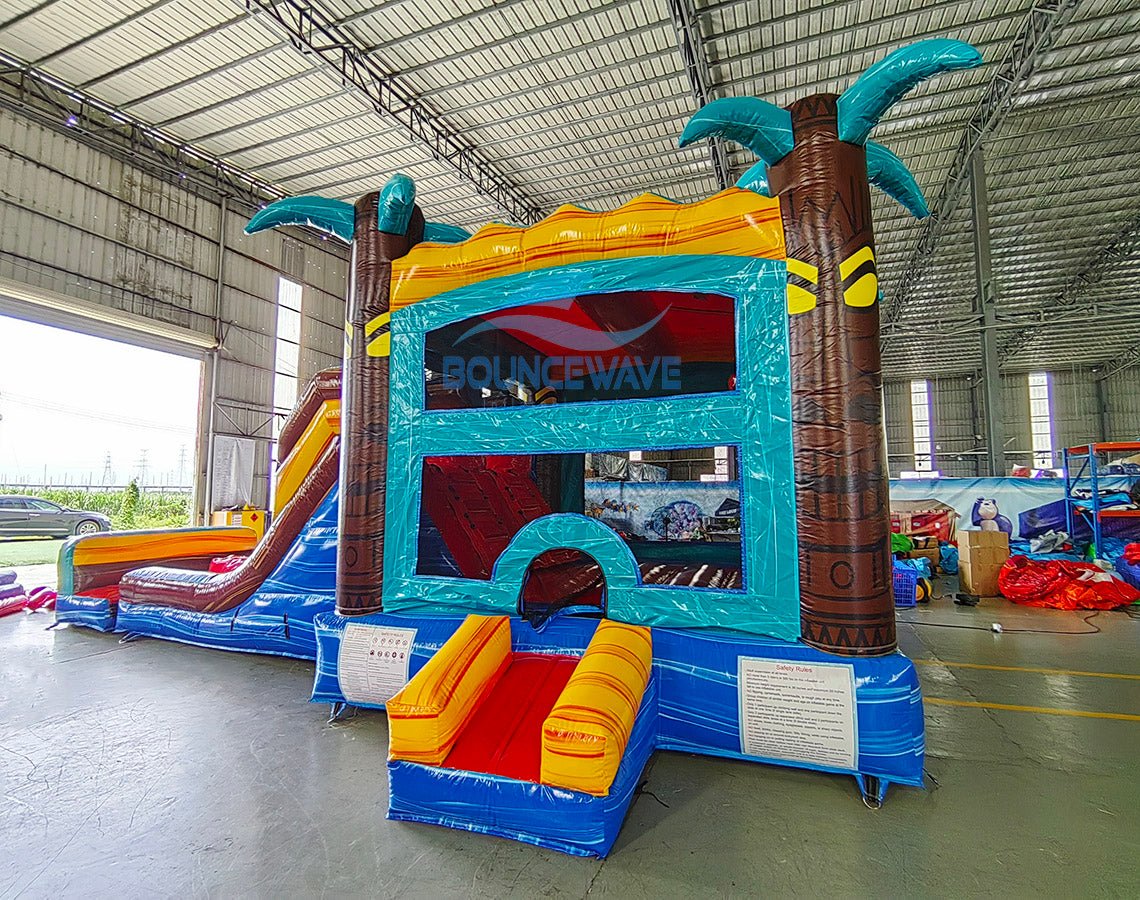 Side view of Tropic Shock Dual Lane Commercial Inflatable Combo (Wet/Dry) showcasing its full length. The inflatable features brown palm tree pillars with turquoise leaves, dual red slides, a turquoise frame, and a colorful base with blue, yellow, and red sections. A large circular splash pool is visible at one end, while the other end shows a yellow bounce house entrance. Set up in a spacious warehouse with metal roofing and various other inflatable products in the background.