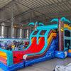 Three-quarter view of Tropic Shock Dual Lane Commercial Inflatable Combo in a spacious warehouse. The inflatable displays turquoise palm trees, dual red slides, brown tree trunk pillars, and a colorful base. A large circular splash pool and a bounce house entrance are visible. Other inflatable products, storage containers, and warehouse materials can be seen in the background, demonstrating its commercial setting.