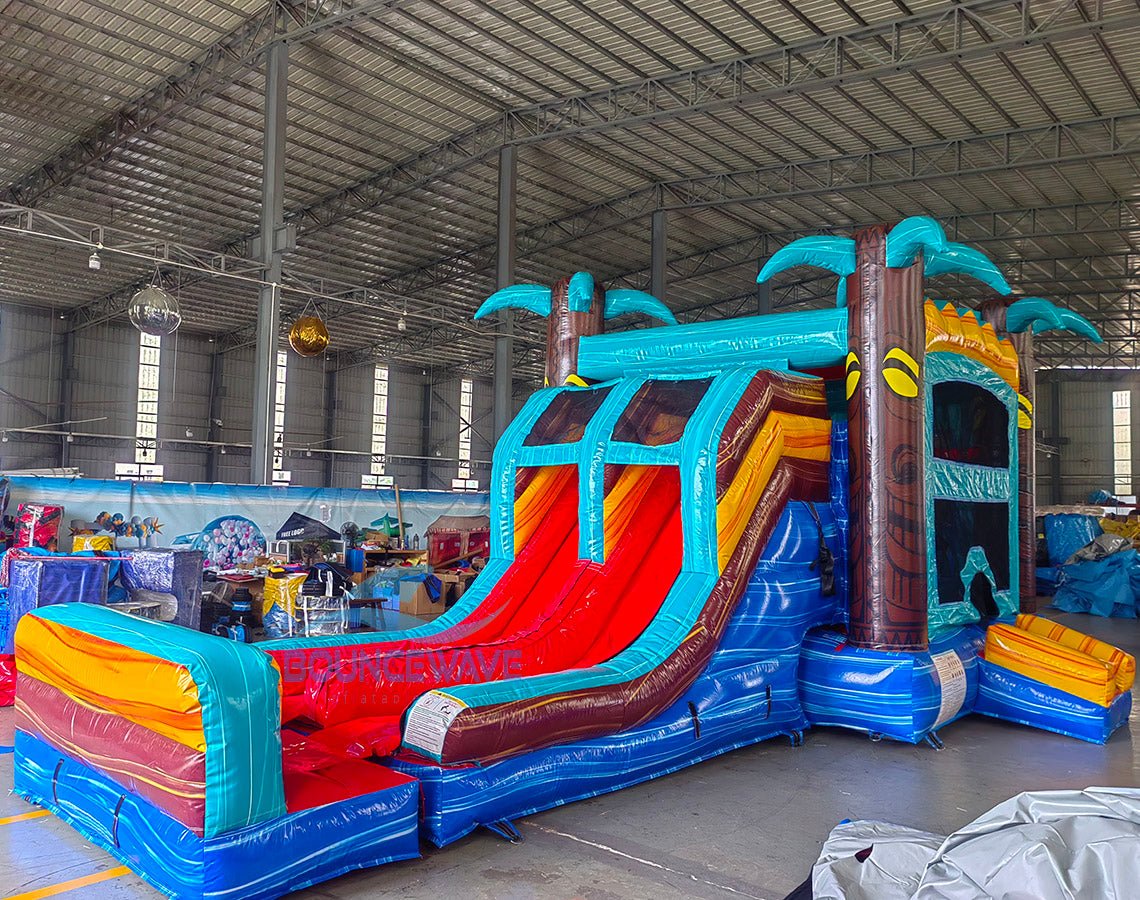 Three-quarter view of Tropic Shock Dual Lane Commercial Inflatable Combo in a spacious warehouse. The inflatable displays turquoise palm trees, dual red slides, brown tree trunk pillars, and a colorful base. A large circular splash pool and a bounce house entrance are visible. Other inflatable products, storage containers, and warehouse materials can be seen in the background, demonstrating its commercial setting.