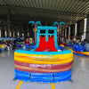 Front view of Tropic Shock Dual Lane Commercial Inflatable Combo set up in a large indoor warehouse. The inflatable features turquoise palm trees, dual red slides, brown tree trunk pillars, and a colorful base with blue, yellow, and red sections. A circular splash pool is prominent in the foreground. Various other inflatable products and warehouse equipment are visible in the background.