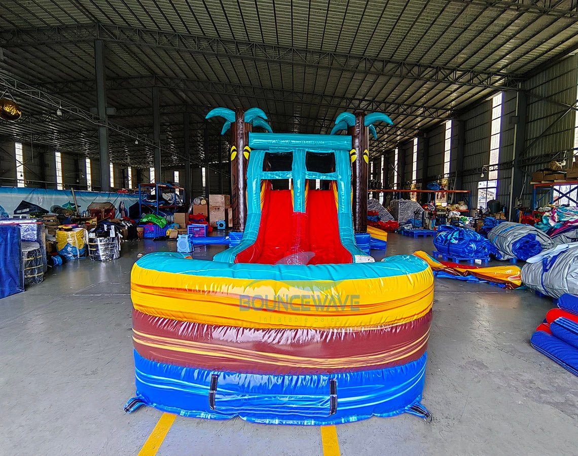 Front view of Tropic Shock Dual Lane Commercial Inflatable Combo set up in a large indoor warehouse. The inflatable features turquoise palm trees, dual red slides, brown tree trunk pillars, and a colorful base with blue, yellow, and red sections. A circular splash pool is prominent in the foreground. Various other inflatable products and warehouse equipment are visible in the background.