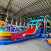 Wide-angle view of Tropic Shock Dual Lane Commercial Inflatable Combo in a warehouse environment. The inflatable features turquoise palm trees, dual red slides, brown tree trunk pillars, and a colorful base with blue, yellow, and red sections. A circular splash pool is prominent. The warehouse setting shows metal roofing, support beams, and various other inflatable products, showcasing its size and commercial application.