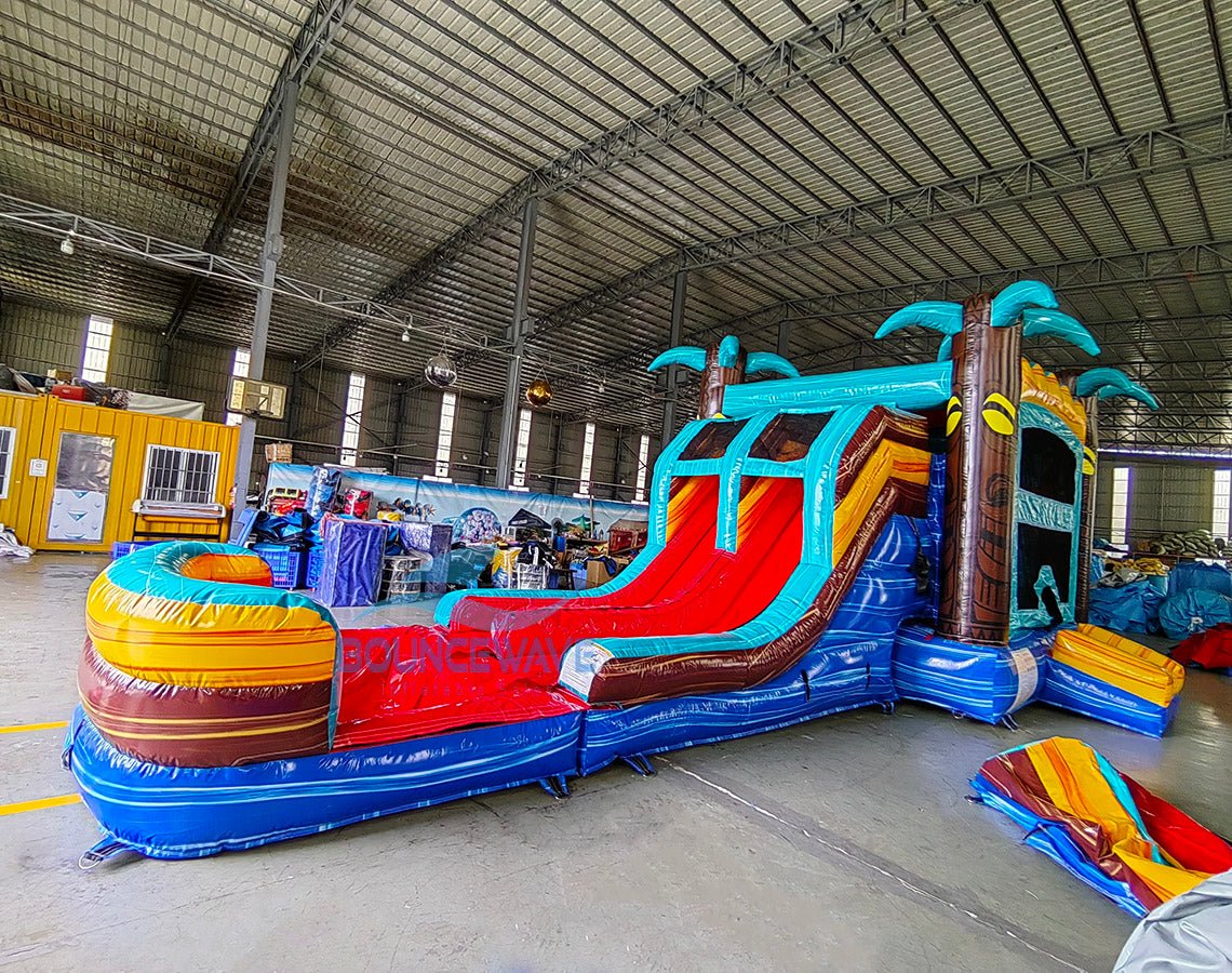 Wide-angle view of Tropic Shock Dual Lane Commercial Inflatable Combo in a warehouse environment. The inflatable features turquoise palm trees, dual red slides, brown tree trunk pillars, and a colorful base with blue, yellow, and red sections. A circular splash pool is prominent. The warehouse setting shows metal roofing, support beams, and various other inflatable products, showcasing its size and commercial application.