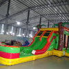 Wide-angle view of Tropical Inferno Dual Lane Commercial Inflatable Combo set up in a spacious warehouse. The structure features dual red slides, brown palm tree trunks with green leaves, and a colorful base. A large green and yellow splash pool area is prominent. The warehouse setting shows metal roofing, support beams, and various other inflatable products, demonstrating its size and commercial application.