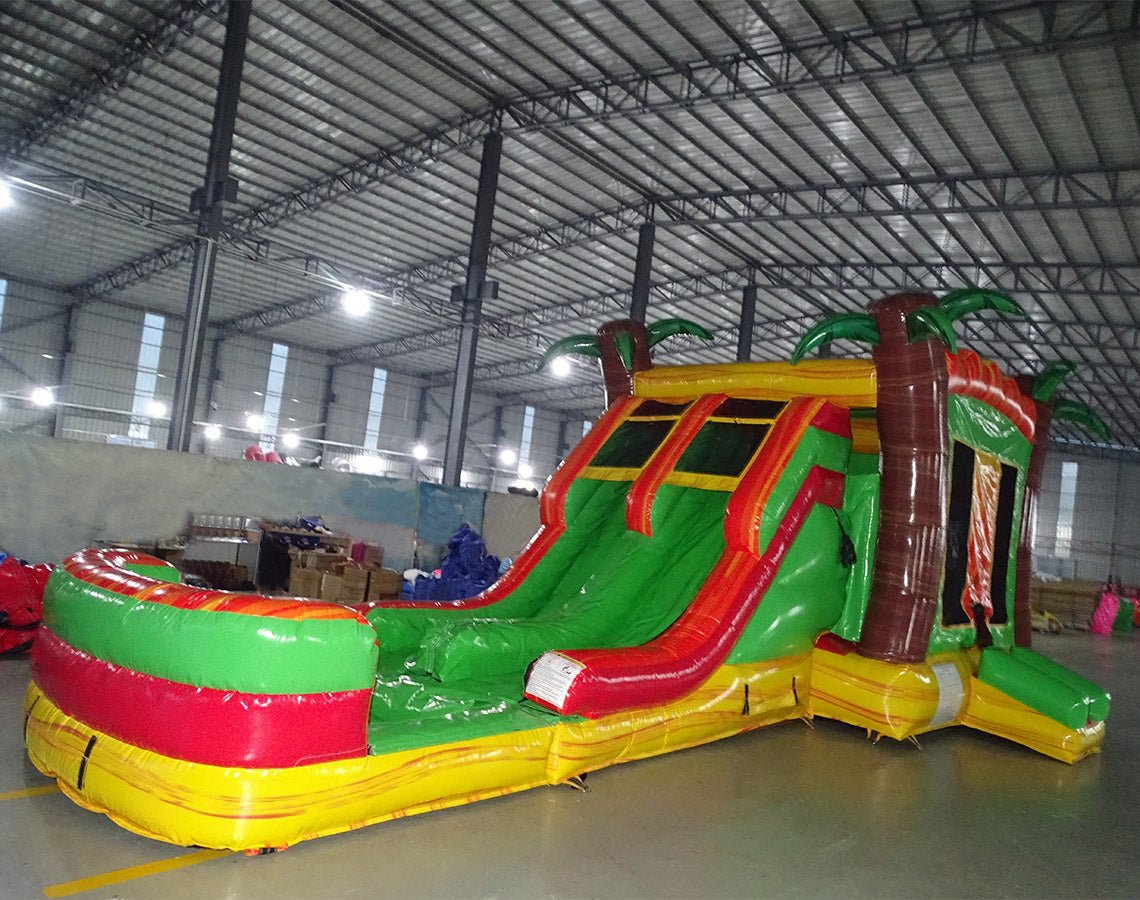 Wide-angle view of Tropical Inferno Dual Lane Commercial Inflatable Combo set up in a spacious warehouse. The structure features dual red slides, brown palm tree trunks with green leaves, and a colorful base. A large green and yellow splash pool area is prominent. The warehouse setting shows metal roofing, support beams, and various other inflatable products, demonstrating its size and commercial application.