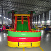 Front view of Tropical Inferno Dual Lane Commercial Inflatable Combo showing dual red slides descending into a green and yellow splash pool. The inflatable features brown palm tree trunks with green leaves, and a yellow top banner. Various other inflatable products and warehouse equipment are visible in the background, showcasing its commercial setting.