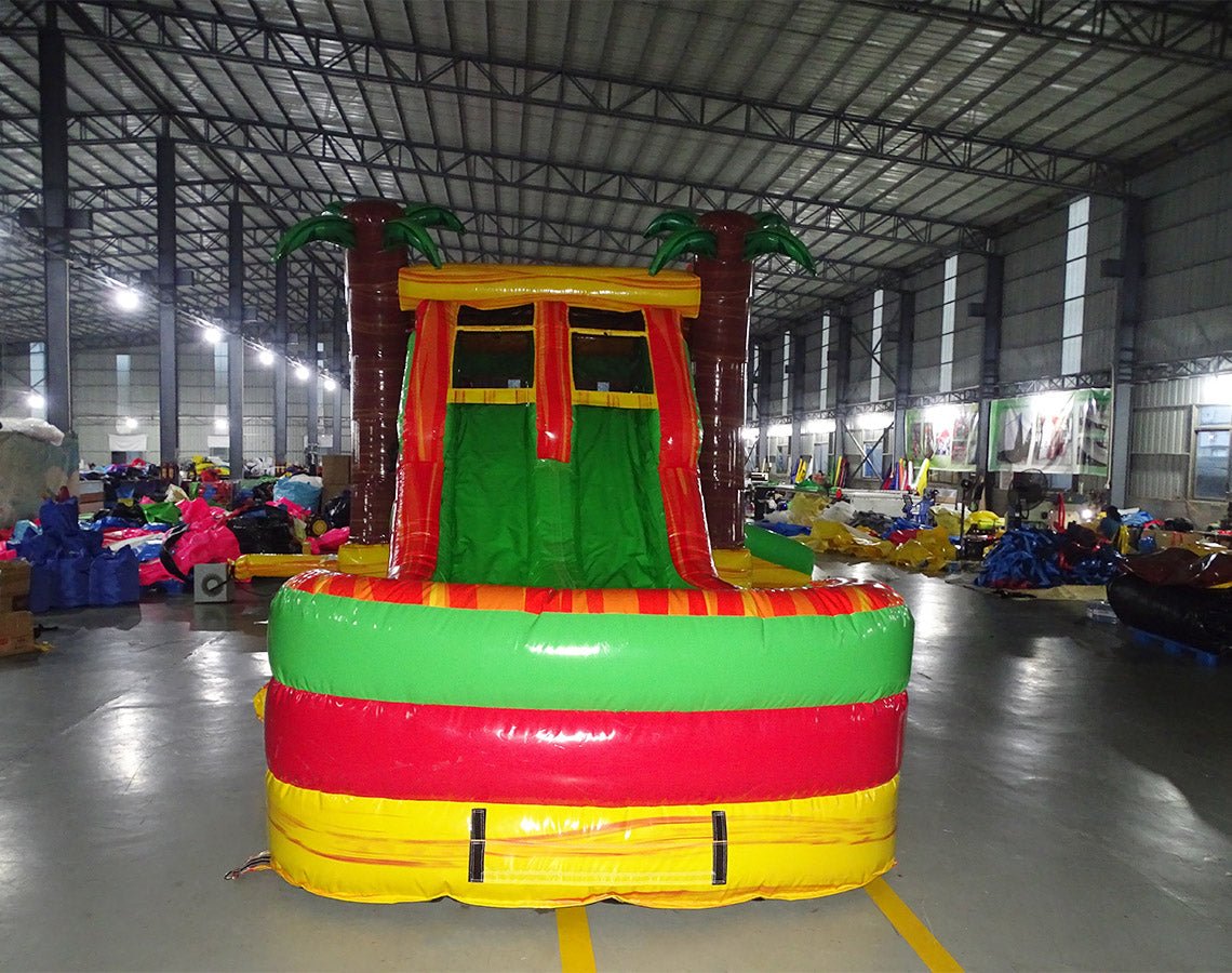 Front view of Tropical Inferno Dual Lane Commercial Inflatable Combo showing dual red slides descending into a green and yellow splash pool. The inflatable features brown palm tree trunks with green leaves, and a yellow top banner. Various other inflatable products and warehouse equipment are visible in the background, showcasing its commercial setting.