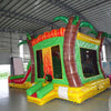 Rear view of Tropical Inferno Dual Lane Commercial Inflatable Combo featuring a vibrant tropical theme. The structure has dual slides in red, emerging from a green base with inflatable palm trees. Brown tree trunks flank the sides, and a colorful splash pool area is visible at the base. Set up in a large indoor warehouse with metal roofing and bright lighting visible.