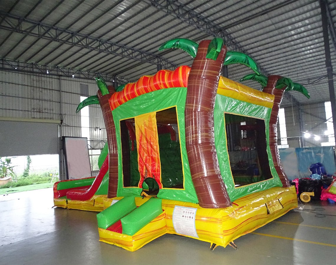 Rear view of Tropical Inferno Dual Lane Commercial Inflatable Combo featuring a vibrant tropical theme. The structure has dual slides in red, emerging from a green base with inflatable palm trees. Brown tree trunks flank the sides, and a colorful splash pool area is visible at the base. Set up in a large indoor warehouse with metal roofing and bright lighting visible.