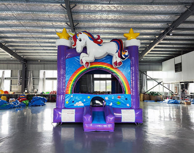 Front view of Unicorn Commercial Grade Bounce House in a spacious warehouse. Purple inflatable with blue sky panels, rainbow arch entrance, and white unicorn figure. Yellow stars top purple towers. Small blue slide at entrance. Warehouse interior visible with metal ceiling, windows, and assorted equipment and materials in background.