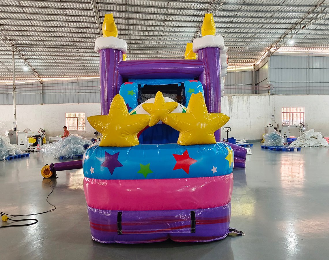 Front view of Unicorn Splash Econo Combo, showcasing the bounce house entrance with rainbow arch, star decorations, and inflatable unicorn figure. The combo includes a slide and play area with bright purple, blue, and pink colors. Set up in an indoor facility with other inflatable products visible.