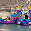 Wide-angle view of Unicorn Splash Econo Combo featuring a bounce house with rainbow arch, star decorations, attached slide, and play area. The inflatable has an eye-catching unicorn figure on top and vibrant purple, blue, pink, and yellow colors. Set up in a spacious indoor warehouse with other equipment and inflatables visible.