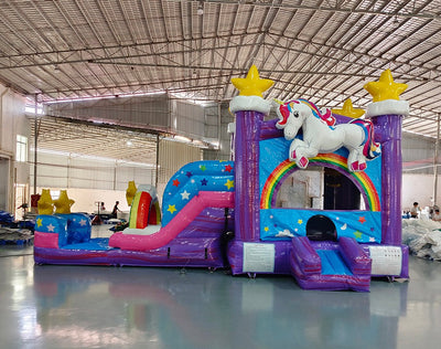 Wide-angle view of Unicorn Splash Econo Combo featuring a bounce house with rainbow arch, star decorations, attached slide, and play area. The inflatable has an eye-catching unicorn figure on top and vibrant purple, blue, pink, and yellow colors. Set up in a spacious indoor warehouse with other equipment and inflatables visible.