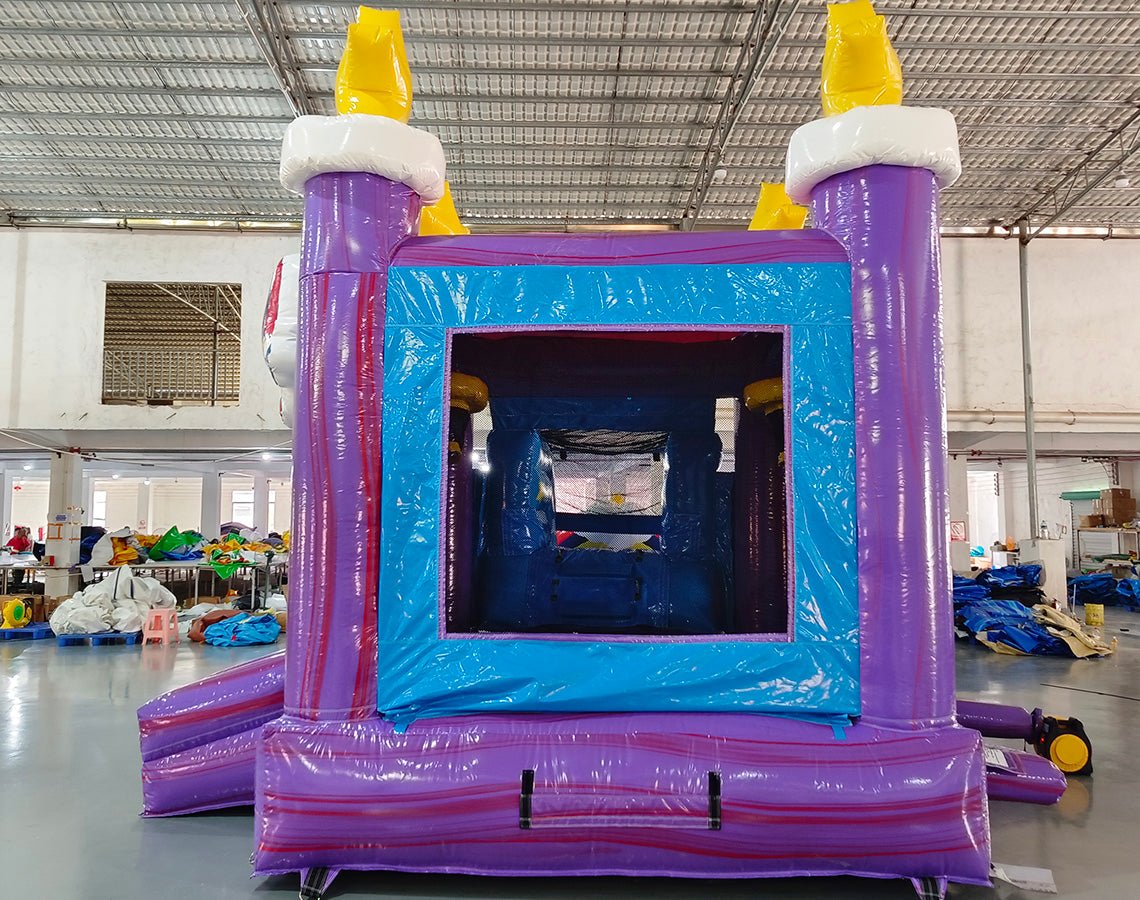 Large inflatable unicorn-themed combo featuring a bounce house with rainbow and star decorations, attached slide, and play area. The structure has vibrant purple, blue, pink, and yellow colors with an inflatable unicorn figure on top. Set up in an indoor warehouse with metal roofing visible.