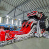 Wide-angle view of UTV Dual Lane Commercial Inflatable Combo set up in a large warehouse. The inflatable features a red and black UTV design with large wheels, dual red slides, and a rock-themed base. The warehouse setting shows metal roofing, support beams, and various other inflatable products and equipment, demonstrating its size and commercial application.