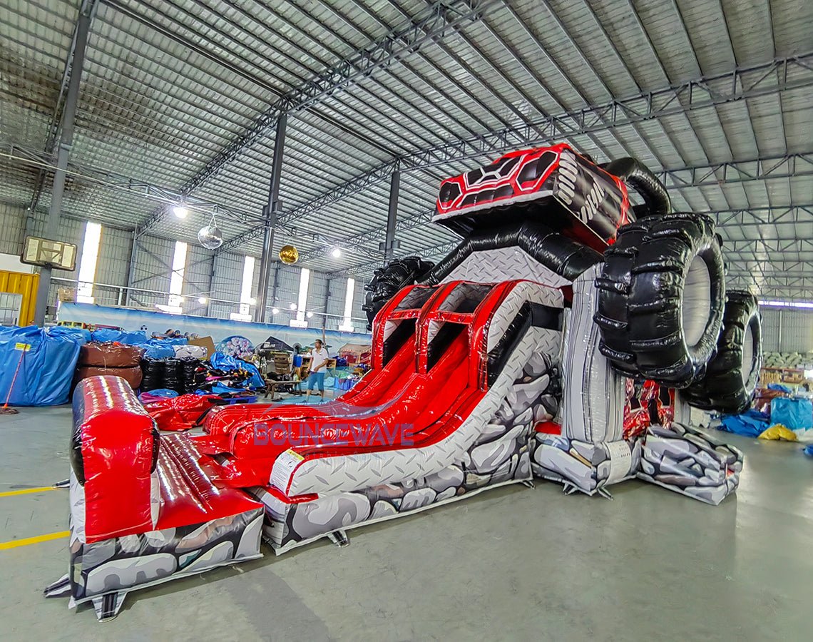 Wide-angle view of UTV Dual Lane Commercial Inflatable Combo set up in a large warehouse. The inflatable features a red and black UTV design with large wheels, dual red slides, and a rock-themed base. The warehouse setting shows metal roofing, support beams, and various other inflatable products and equipment, demonstrating its size and commercial application.