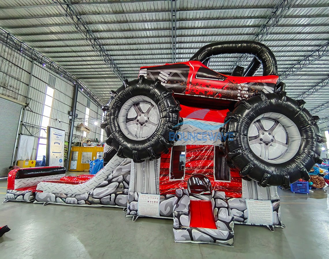 Side view of UTV Dual Lane Commercial Inflatable Combo featuring a large red and black inflatable UTV (Utility Task Vehicle) structure. The combo includes dual red slides emerging from the UTV body, large black inflatable wheels, and a rock-themed base. Set up in a spacious warehouse with metal roofing and bright lighting visible.