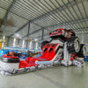 Side view of UTV Dual Lane Commercial Inflatable Combo showing the full length of the inflatable. The structure features a red and black UTV design with large black wheels, dual red slides, and a rock-patterned base. Various other inflatable products and warehouse equipment are visible in the background, showcasing its commercial setting.