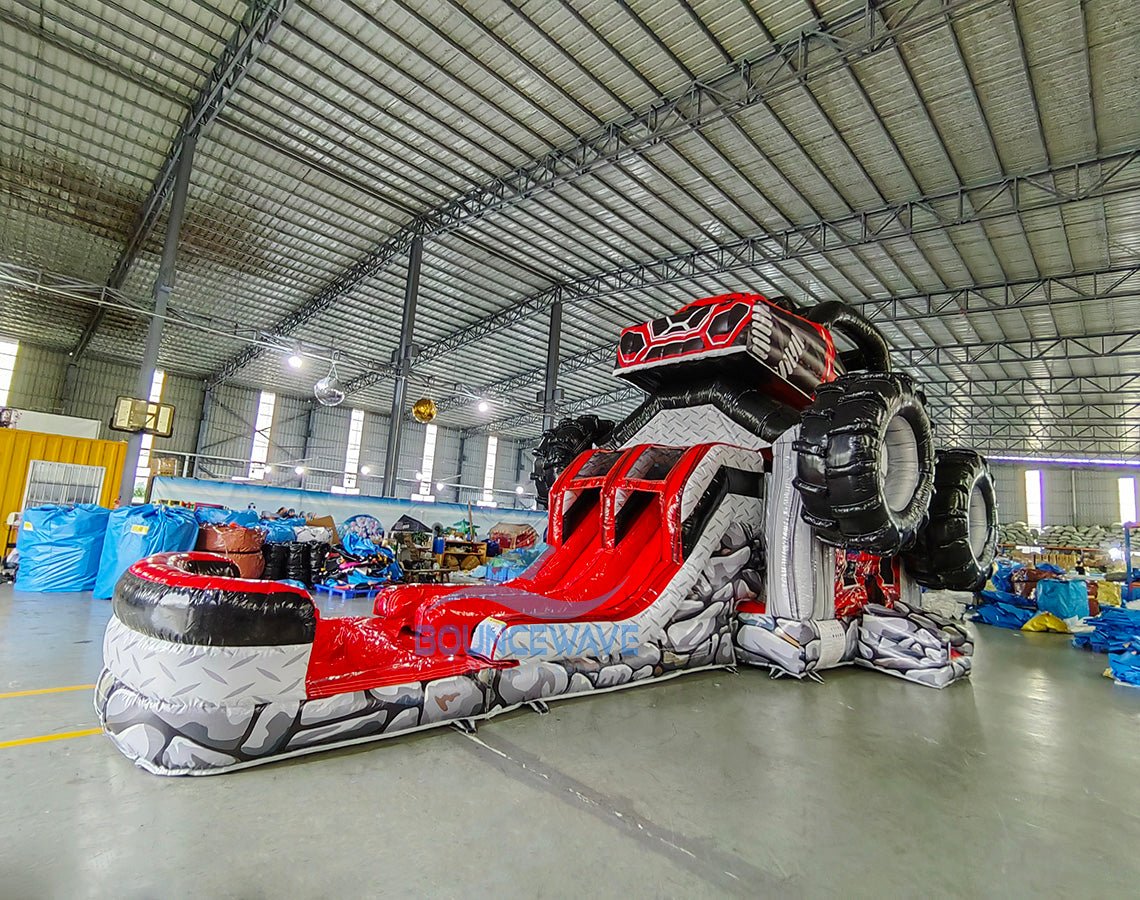 Side view of UTV Dual Lane Commercial Inflatable Combo showing the full length of the inflatable. The structure features a red and black UTV design with large black wheels, dual red slides, and a rock-patterned base. Various other inflatable products and warehouse equipment are visible in the background, showcasing its commercial setting.