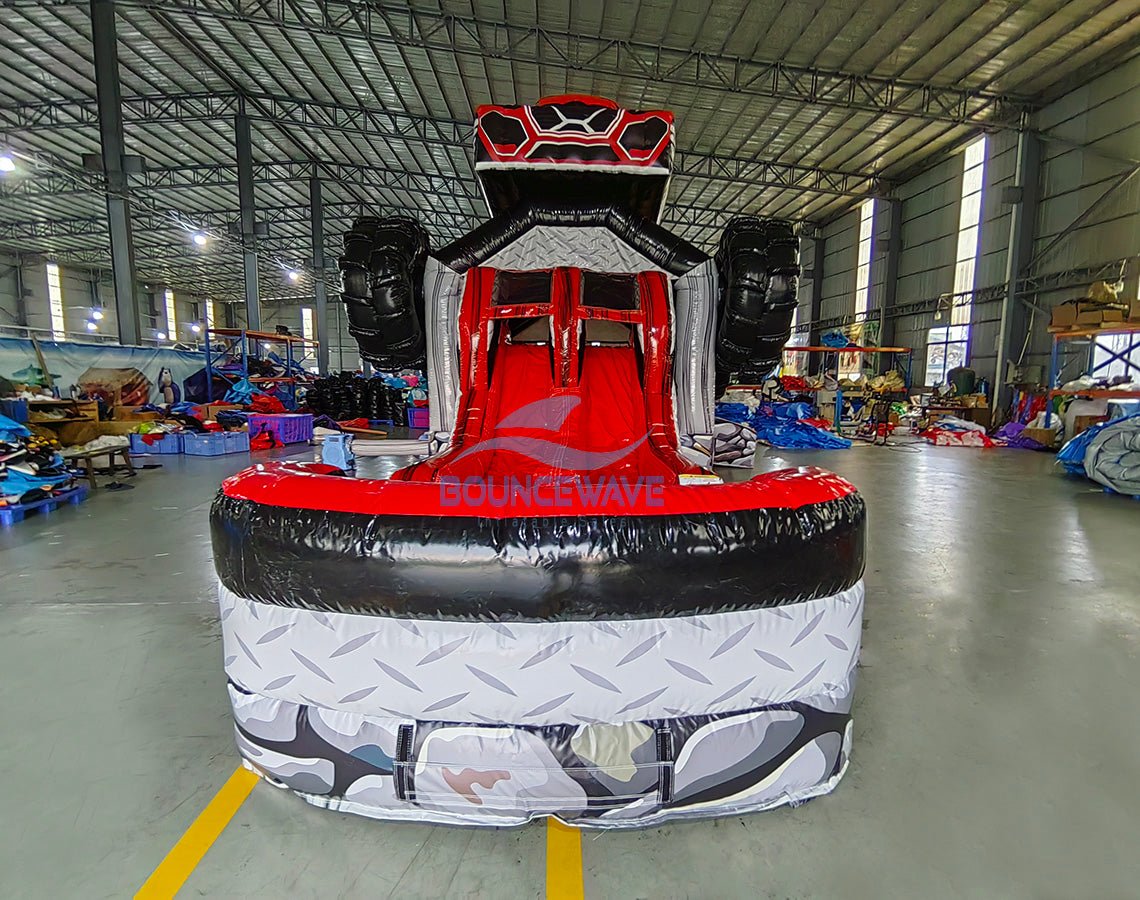 Front view of UTV Dual Lane Commercial Inflatable Combo (Wet/Dry) featuring a large red and black inflatable UTV (Utility Task Vehicle) structure. The combo includes dual red slides emerging from the UTV body, large black inflatable wheels, and a rock-themed base with a diamond plate pattern. The "BOUNCEWAVE" logo is visible on the front. Set up in a spacious warehouse with metal roofing, bright lighting, and various other inflatable products visible in the background.