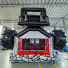 Rear view of UTV Dual Lane Commercial Inflatable Combo (Wet/Dry) showing the full length of the inflatable. The structure features a red and black UTV design with large black wheels, dual red slides, and a rock-patterned base with a diamond plate design. The "BOUNCEWAVE" logo is prominently displayed. Set in a large warehouse with metal roofing, support beams, and various other inflatable products and equipment visible, demonstrating its size and commercial application.