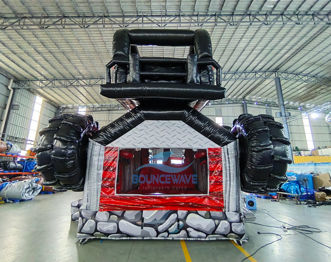 Rear view of UTV Dual Lane Commercial Inflatable Combo (Wet/Dry) showing the full length of the inflatable. The structure features a red and black UTV design with large black wheels, dual red slides, and a rock-patterned base with a diamond plate design. The "BOUNCEWAVE" logo is prominently displayed. Set in a large warehouse with metal roofing, support beams, and various other inflatable products and equipment visible, demonstrating its size and commercial application.