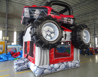 Wide-angle view of UTV Trail Boss Commercial Grade Bounce House in a spacious warehouse. The inflatable off-road vehicle design features oversized black tires, a red and black body, and a detailed roll cage. The red bounce area entrance is visible beneath the vehicle. The warehouse setting shows metal roofing, floor markings, and various other inflatable products, demonstrating its size and commercial application.